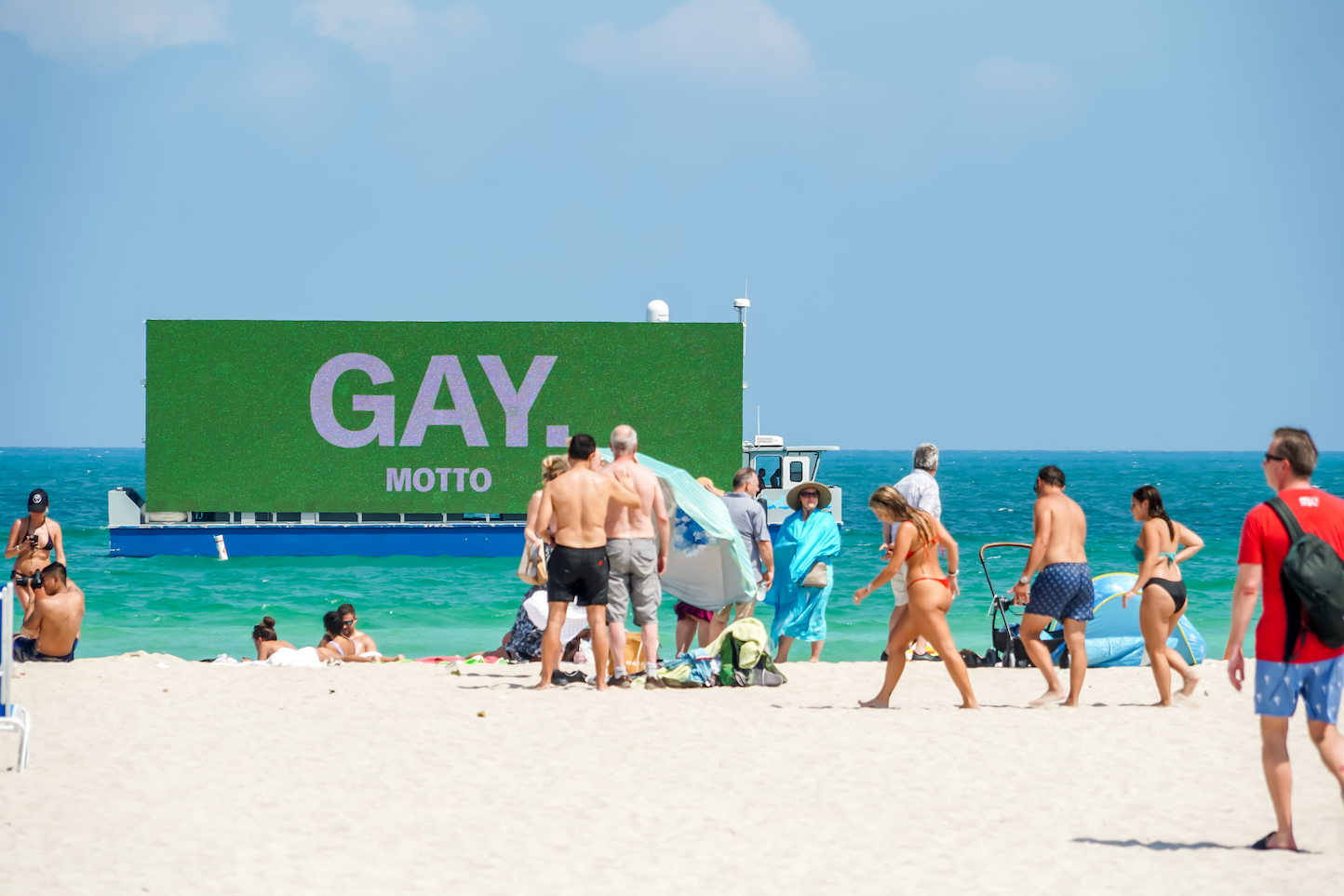 A boat with a digital billboard of the Motto campaign reading GAY seen in the distance behind a group of people at a beach